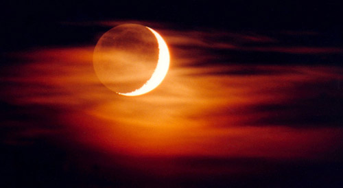 Un fin voile nuageux masque le croissant lunaire surexposé, mais laisse voir la lumière cendrée, cette pâle lueur qui suffit à éclairer la surface lunaire normalement plongée dans l’obscurité. On y distingue faiblement mers et cratères.